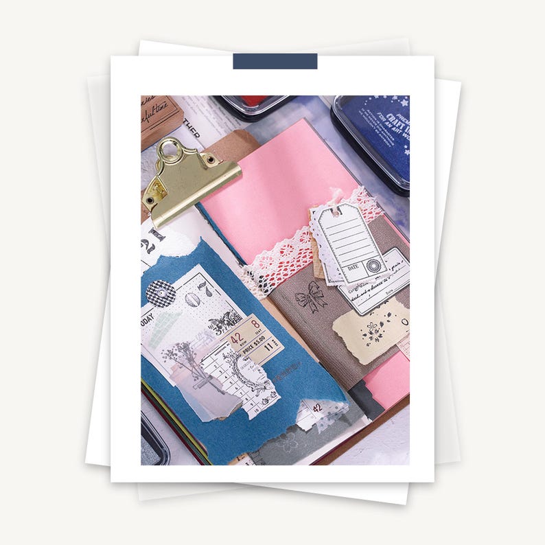 May include: A close-up of a scrapbook page with various decorative elements, including paper scraps, stickers, stamps, and tags. The page is decorated with a pink and blue color scheme and features a gold binder clip.