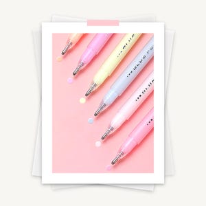 May include: A collection of pastel-colored highlighters on a pink background. The highlighters are in shades of pink, yellow, and blue. The text "...BLUE FE..." is visible on the side of the highlighters. Small, colorful dots are scattered around the highlighters.