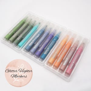 Peut inclure: Ensemble de surligneurs à paillettes dans un étui en plastique transparent. Les marqueurs sont de différentes couleurs, dont le vert, le bleu, le violet, l'orange et le rose. Le texte "Glitter Highter Markers" est sur un cercle rose.