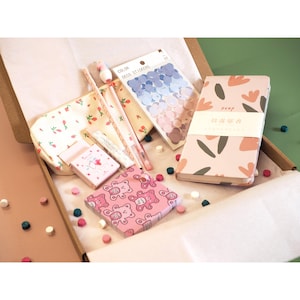 May include: A stationery set in a cardboard box, featuring a pink floral notebook, stickers, pens, a small box, and a pouch with a strawberry pattern. The set includes a pink box with a teddy bear pattern and small decorative beads.