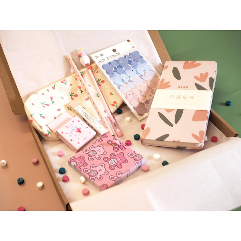 May include: A stationery set in a cardboard box, featuring a pink floral notebook, stickers, pens, a small box, and a pouch with a strawberry pattern. The set includes a pink box with a teddy bear pattern and small decorative beads.