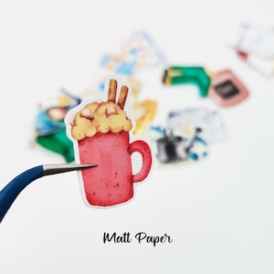 May include: A watercolor sticker of a red mug filled with a creamy beverage, topped with whipped cream and cinnamon sticks. The sticker is held by a blue-handled tool. The text "Matt Paper" is at the bottom.