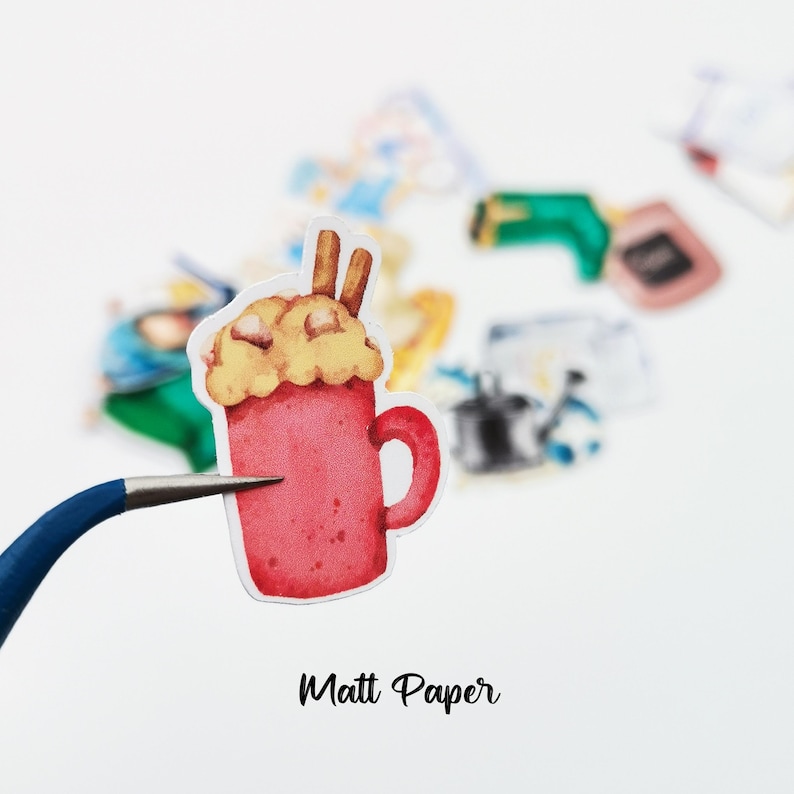 May include: A watercolor sticker of a red mug filled with a creamy beverage, topped with whipped cream and cinnamon sticks. The sticker is held by a blue-handled tool. The text "Matt Paper" is at the bottom.