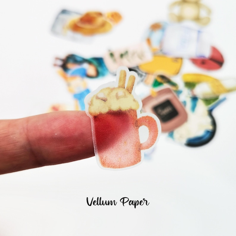 May include: A sticker of a red mug with a cream-colored topping and two sticks. The sticker is held by a finger. The text "Vellum Paper" is at the bottom of the image. Other stickers are in the background.