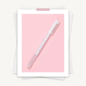 May include: A white highlighter pen with a clear tip and a silver clip, set against a soft pink background. The pen has subtle text along its side. The image is presented on a white paper background.
