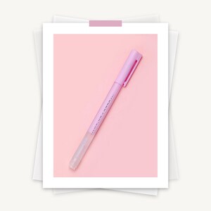 May include: A light purple pen with a matching cap and clip, set against a pink background. The pen has a clear tip and a small amount of text printed on the side. The pen is taped to a white photo frame.