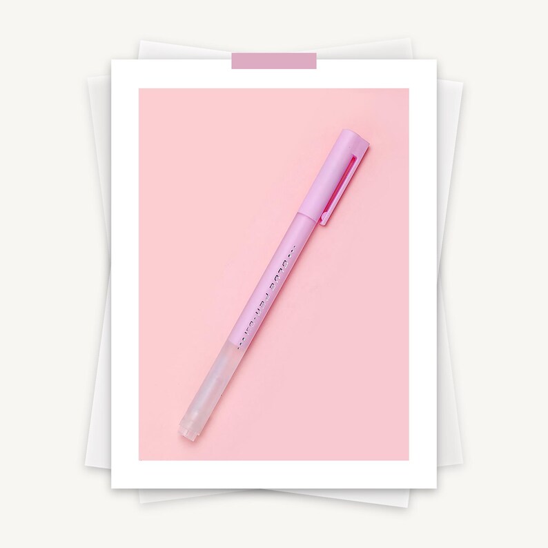 May include: A light purple pen with a matching cap and clip, set against a pink background. The pen has a clear tip and a small amount of text printed on the side. The pen is taped to a white photo frame.