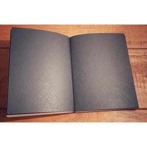 May include: A black notebook with a textured cover is open to a blank page. The notebook is lying flat on a wooden surface.