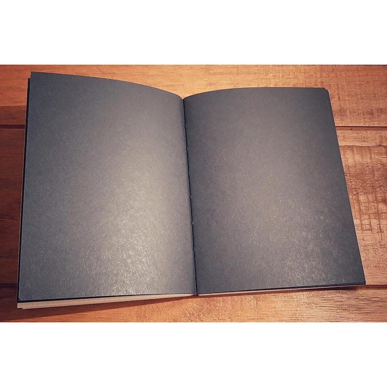 May include: A black notebook with a textured cover is open to a blank page. The notebook is lying flat on a wooden surface.