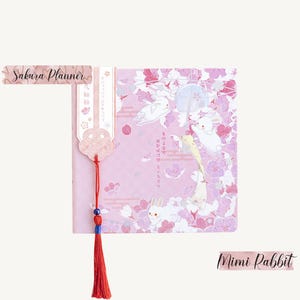 Puede incluir: Una agenda rosa con un diseño floral con conejos blancos y flores de cerezo. La agenda tiene un marcapáginas de borla roja con una cuenta azul. El texto "Sakura Planner" está en la parte superior de la agenda y el texto "Mimi Rabbit" está en la parte inferior.