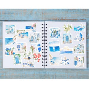 May include: A collection of watercolor stickers featuring Greek island scenes. The stickers depict white buildings with blue accents, coastal landscapes, and floral arrangements. The stickers are arranged on a white spiral-bound notebook.