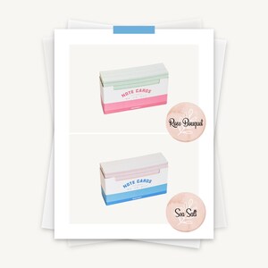 May include: Two boxes of note cards, one with a pink and green design and the text "Note Cards" and the other with a blue and white design and the text "Note Cards". The boxes are on a white background with a pink circle with the text "Rose Bouquet" and a blue circle with the text "Sea Salt".