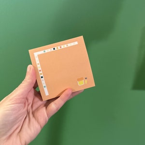 May include: A peach-colored sticky note with a white border and a small illustration of a jar of honey and a paintbrush. The border is decorated with a row of small, colorful squares.