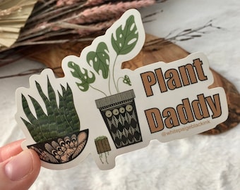May include: A sticker with a green monstera plant, a snake plant, and the text "Plant Daddy" in brown lettering. The sticker also includes the text "@whitepaigeblackink" in smaller lettering.