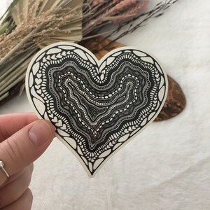 May include: A black and white heart-shaped sticker with a detailed, intricate design. The design features a series of concentric circles and wavy lines, creating a mesmerizing pattern.