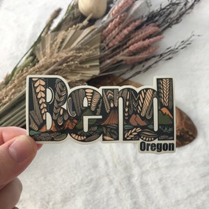 May include: A sticker with a stylized design of the word "Bend" in black and brown with a green and brown mountain scene. The word "Oregon" is printed below the word "Bend".