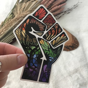 May include: A colorful sticker of a raised fist with a rainbow pattern. The fist is outlined in black and filled with intricate designs in shades of red, orange, yellow, green, blue, and purple.