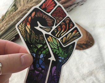 May include: A colorful sticker of a raised fist with a rainbow pattern. The fist is outlined in black and filled with intricate designs in shades of red, orange, yellow, green, blue, and purple.