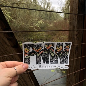 Puede incluir: Una pegatina con las letras "PNW" en una fuente estilizada, con una cordillera y l&iacute;neas onduladas en el fondo. El texto "Pacific Northwest" est&aacute; debajo de las letras.