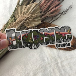 May include: A sticker with the word "Eugene" in a stylized font with a mountain range and flowers in the background. The text "Oregon" is below the word Eugene.