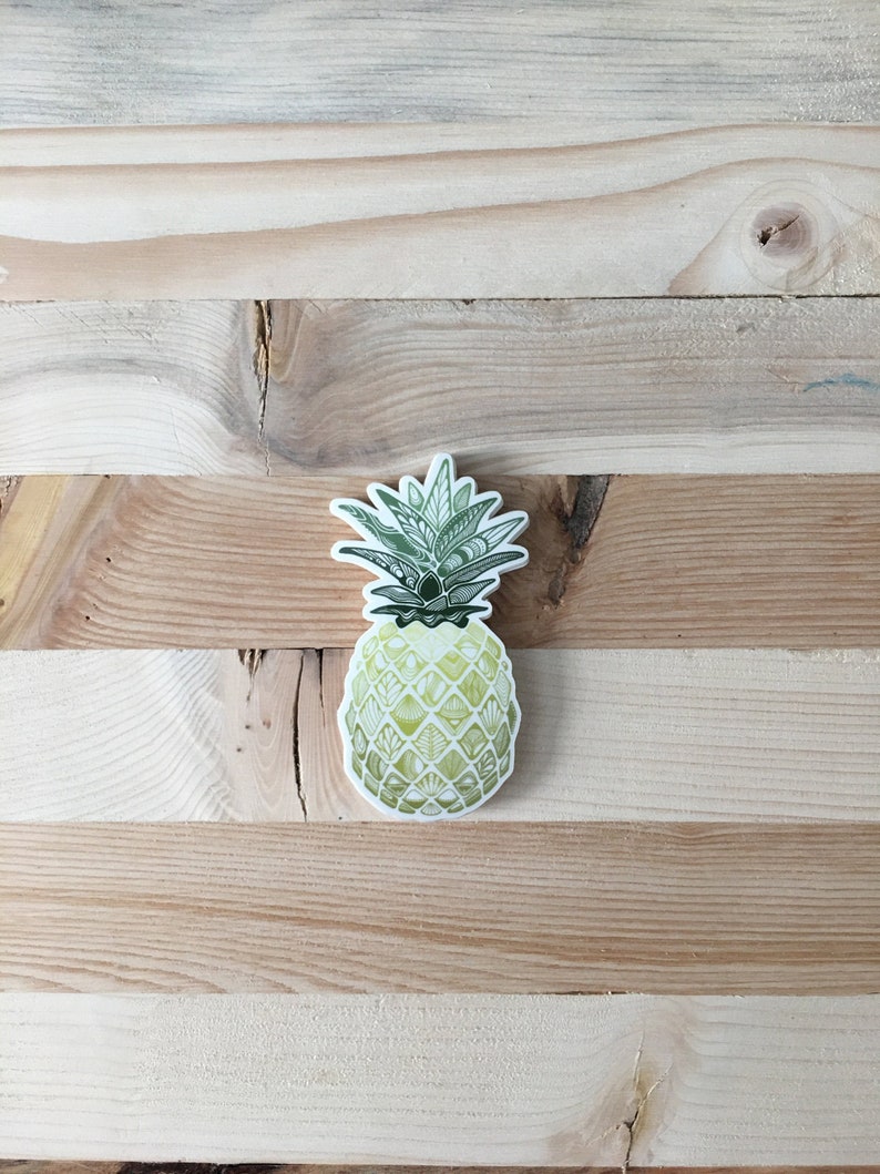 May include: A green and yellow pineapple sticker with a detailed design. The pineapple is on a light brown wooden background.