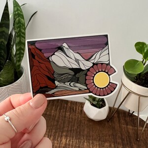 May include: A sticker featuring a colorful illustration of a mountain range with a sun setting behind it. The mountains are white with black outlines, and the sky is a gradient of purple, pink, and orange. The sun is yellow with a red and pink flower around it.