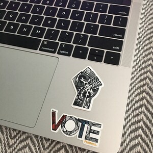 May include: A silver laptop computer with a black keyboard and a trackpad. Two stickers are on the laptop. One sticker is a black and white stylized fist with intricate details. The other sticker is a colorful stylized word "VOTE" with the text "Your Vote Matters." below it.