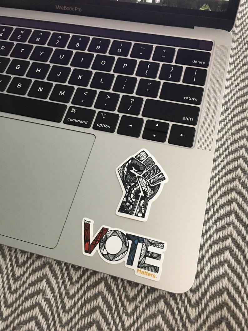 May include: A silver laptop computer with a black keyboard and a trackpad. Two stickers are on the laptop. One sticker is a black and white stylized fist with intricate details. The other sticker is a colorful stylized word "VOTE" with the text "Your Vote Matters." below it.