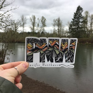 Puede incluir: Una pegatina con las letras "PNW" en una fuente estilizada, con un dise&ntilde;o de cordillera en tonos naranja, amarillo, verde y gris. El texto "Pacific Northwest" est&aacute; escrito debajo de las letras.