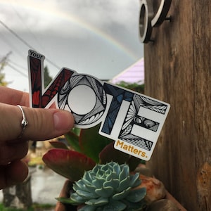 May include: A white sticker with the word "VOTE" in black outlined letters with a colorful, intricate design. The sticker also says "Your Vote Matters." in gold lettering.