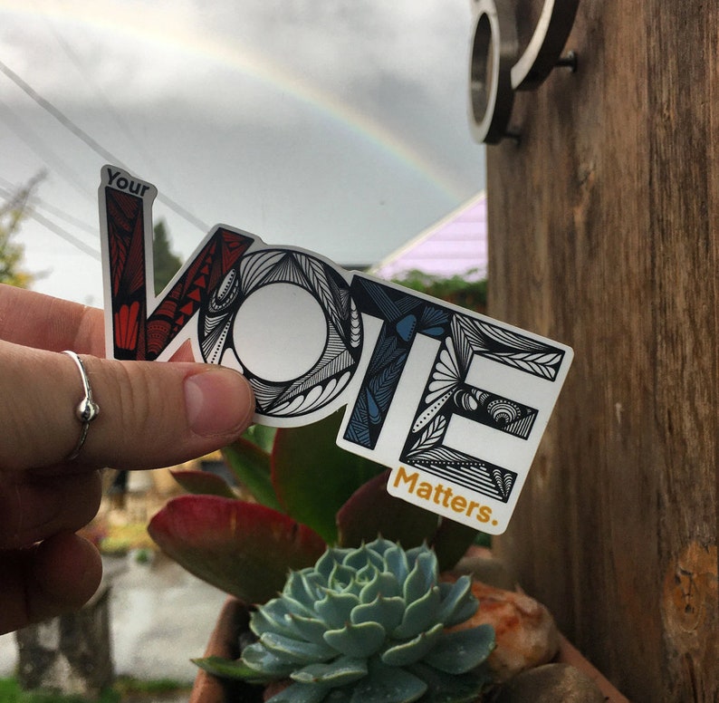 May include: A white sticker with the word "VOTE" in black outlined letters with a colorful, intricate design. The sticker also says "Your Vote Matters." in gold lettering.