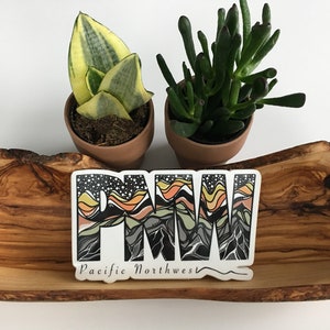 Puede incluir: Una pegatina blanca con dise&ntilde;os de monta&ntilde;a negros, naranja, amarillos y verdes que deletrean "PNW" y el texto "Pacific Northwest". La pegatina est&aacute; sobre una superficie de madera con dos plantas en maceta.