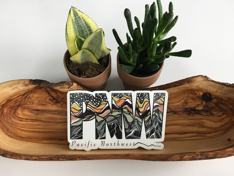 Puede incluir: Una pegatina blanca con dise&ntilde;os de monta&ntilde;a negros, naranja, amarillos y verdes que deletrean "PNW" y el texto "Pacific Northwest". La pegatina est&aacute; sobre una superficie de madera con dos plantas en maceta.