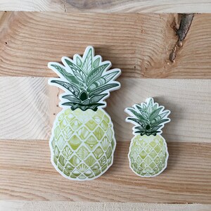 May include: Two pineapple stickers with a green and yellow patterned design. The larger sticker is on the left and the smaller sticker is on the right.