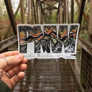 Puede incluir: Una pegatina con las letras "PNW" en una fuente estilizada, con un dise&ntilde;o de cordillera en tonos naranja, amarillo, verde y gris. El texto "Pacific Northwest" est&aacute; debajo de las letras.