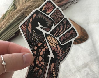 May include: A black and brown patterned sticker of a raised fist. The fist is stylized with intricate details and a variety of shapes.