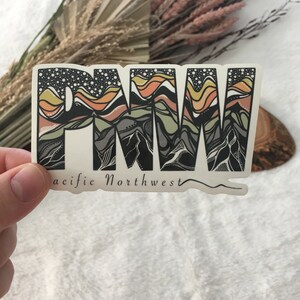 Puede incluir: Una pegatina con un gr&aacute;fico estilizado de las letras "PNW" en negro, blanco y tonos de marr&oacute;n y amarillo. Las letras est&aacute;n rellenas con un dise&ntilde;o de cordillera. El texto "Pacific Northwest" est&aacute; escrito debajo de las letras.