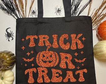 May include: A black canvas tote bag featuring the orange glitter text TRICK OR TREAT, with the letter O replaced by a jack-o'-lantern pumpkin graphic. The design includes small orange glitter stars and bat silhouettes surrounding the text.