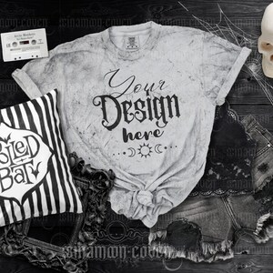 Könnte beinhalten: Ein weißes T-Shirt mit einem Used-Look und dem Text "Your Design Here" in Schwarz. Das Shirt liegt auf einer dunklen Holzoberfläche mit einem schwarz-weiß gestreiften Kissen, einem Schädel und einer Jeans-Shorts.