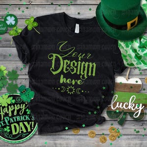 Puede incluir: Una camiseta negra con el texto "Your Design Here" en letras verdes. La camiseta está rodeada de tréboles, monedas de oro y otras decoraciones del Día de San Patricio.
