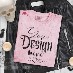 Puede incluir: Camiseta rosa con un aspecto desgastado y el texto "Your Design here" en fuente g&oacute;tica negra. La camiseta est&aacute; sobre una chaqueta de cuero negra con un cr&aacute;neo y velas en el fondo.