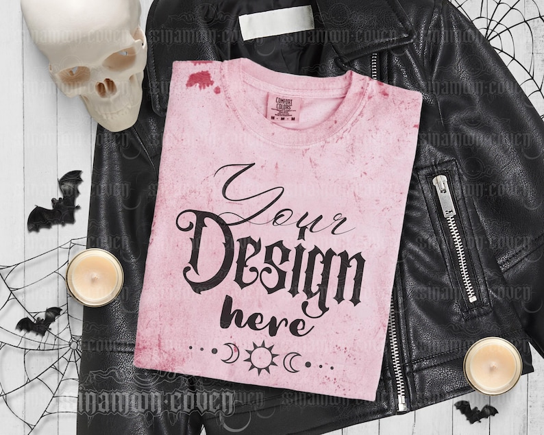 Puede incluir: Camiseta rosa con un aspecto desgastado y el texto "Your Design here" en fuente g&oacute;tica negra. La camiseta est&aacute; sobre una chaqueta de cuero negra con un cr&aacute;neo y velas en el fondo.