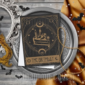 May include: A black and gold birthday card with a cake illustration and the text "THE BIRTHDAY". The card is on a silver tray with a gold chain and a gold and silver ornate frame. Decorative bats are scattered around the scene.