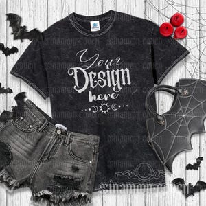 Könnte beinhalten: Ein schwarzes T-Shirt mit einem Used-Look und dem Text "Your Design Here" in Weiß. Das Shirt liegt auf einem weißen Holzgrund mit einer schwarzen Jeans-Shorts und einer schwarzen Fledermaus-Handtasche mit Spinnennetz-Design.