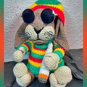 Könnte beinhalten: Ein gehäkeltes braunes Kaninchen mit einem rot, gelb und grün gestreiften Hut, runden Sonnenbrillen und einem passenden gestreiften Hemd. Das Kaninchen hält eine gehäkelte Zigarette. Der Hintergrund ist ein neutrales Grau.