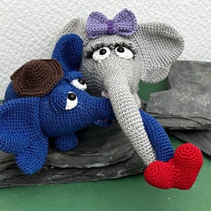 Könnte beinhalten: Zwei gehäkelte Elefantenpuppen. Einer ist grau mit einer lila Schleife, der andere ist blau mit einem braunen Hut. Der graue Elefant hat lange Wimpern und einen Rüssel, der den blauen Elefanten berührt. Der blaue Elefant hat einen roten herzförmigen Fuß.