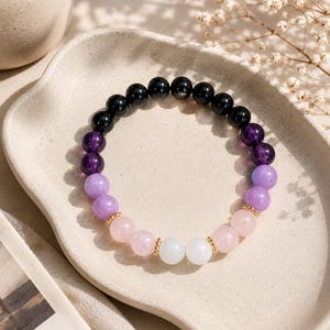 May include: A beaded bracelet featuring a gradient of smooth, round stones in black, deep purple, light lavender, soft pink, and white. The beads are arranged in a circular pattern and accented with small, textured gold-toned metal spacers.