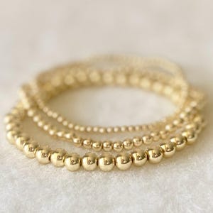 May include: Three gold beaded bracelets of varying sizes are stacked on a white surface. The bracelets have a shiny, metallic finish and are arranged in a slightly curved pattern, showcasing the different bead sizes.
