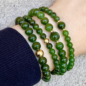 May include: Four jade green beaded bracelets with gold accents are stacked on a wrist. The bracelets feature round, polished beads of varying sizes. The gold accents are interspersed throughout the bracelets, adding a touch of elegance to the jewellery.