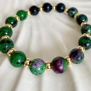 May include: A bracelet with green and purple gemstone beads and gold-coloured beads. The bracelet is presented on a white, textured surface. The gemstones have a speckled appearance. The beads are likely around 8mm in diameter.